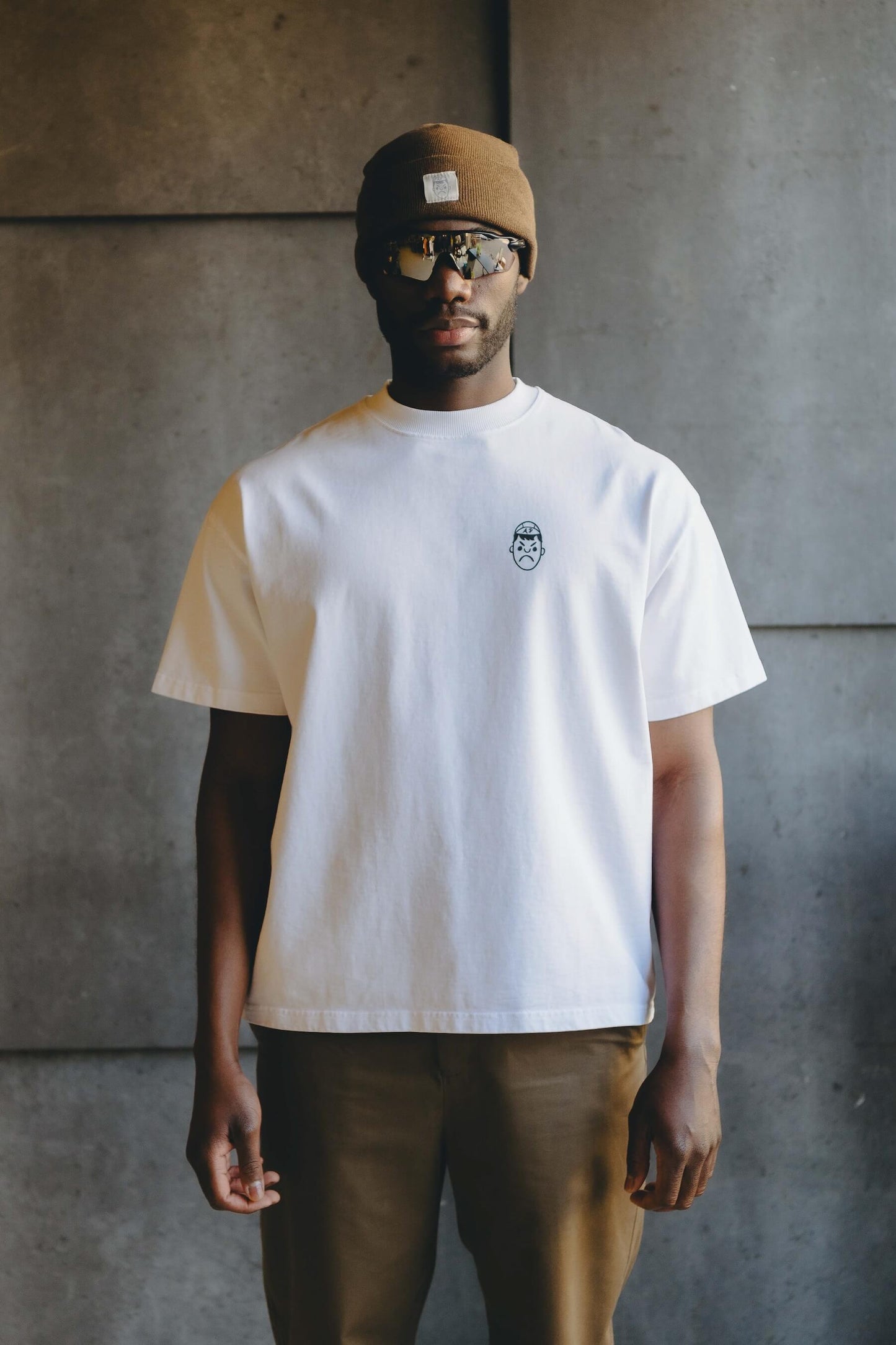 A man wearing a heavy overiszed white t-shirt, Oakley sunglasses and brown beanie hat.