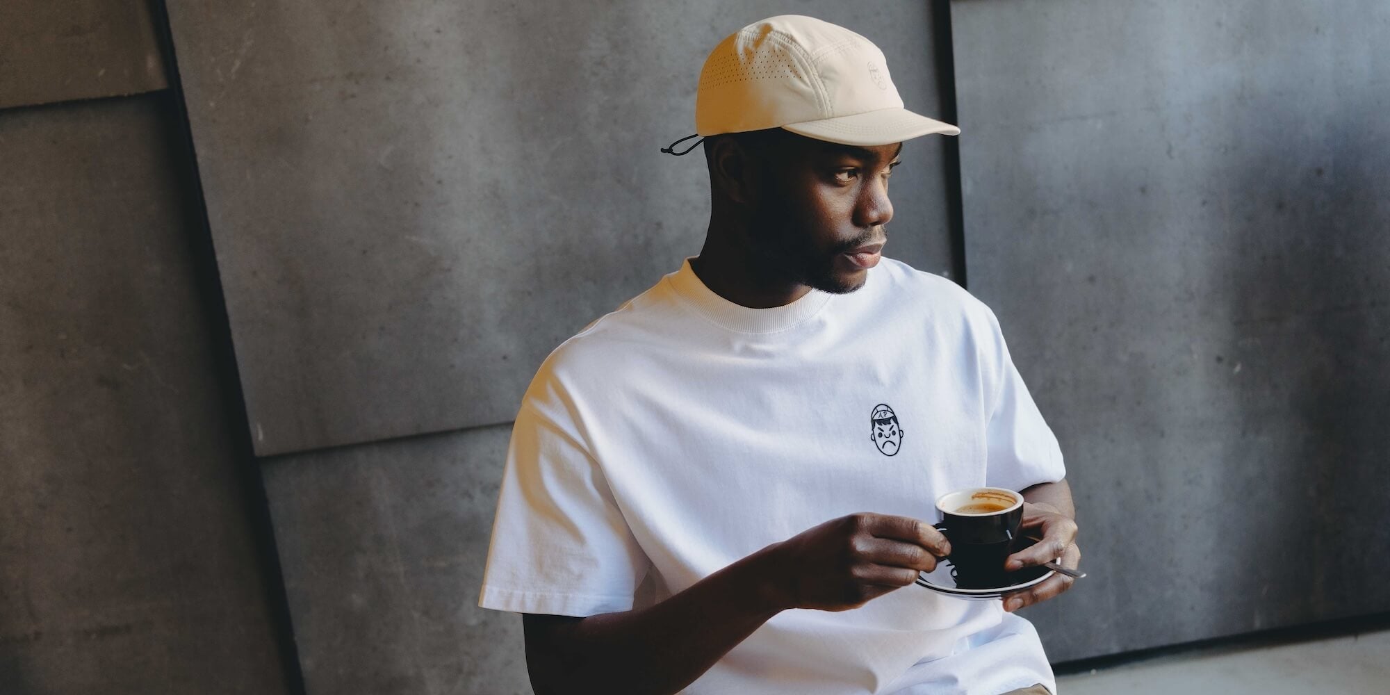 A man drinking a coffee whilst wearing a running cap and oversized white t-shirt